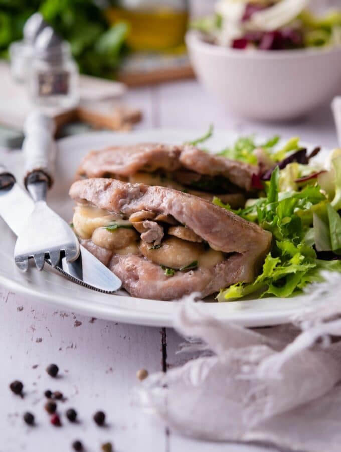 Seared pork chops stuffed with swiss cheese and mushrooms on a white plate with garden salad. A fork and knife are on the plate and a bowl of salad can be seen in the back.