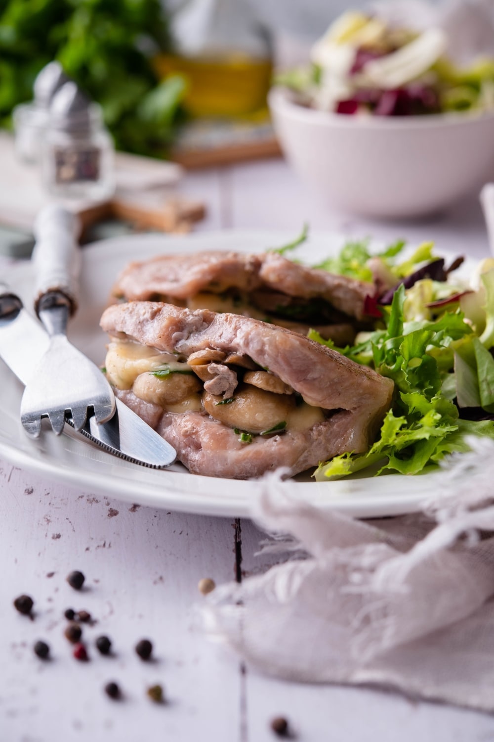 Seared pork chops stuffed with swiss cheese and mushrooms on a white plate with garden salad. A fork and knife are on the plate and a bowl of salad can be seen in the back.