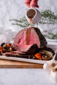 A sliced medium rare eye of round roast on a white rectangle plate with mushrooms and carrots. Gravy is being poured onto the roast from a small ceramic pitcher.