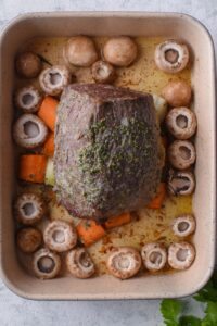 Partially cooked eye of round steak on a ceramic baking tray with mushrooms, carrots, and onions.