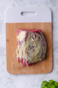 A raw eye of round steak coated in seasoned butter on a wooden cutting board with a white handle.