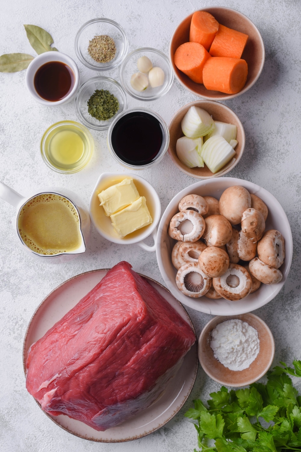 A plate of raw eye of round steak, ceramic bowls of mushrooms, chopped carrots, chopped onion, and butter, and smaller bowls of herbs, garlic, wine, beef stock, Worcestershire sauce, and cornstarch.