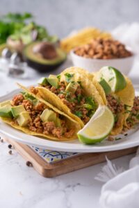 Crunchy shell tacos stuffed with seasoned ground turkey, avocados, and cilantro on a plate with a lime wedge. Behind is a bowl of seasoned ground turkey meat and a sliced avocado.