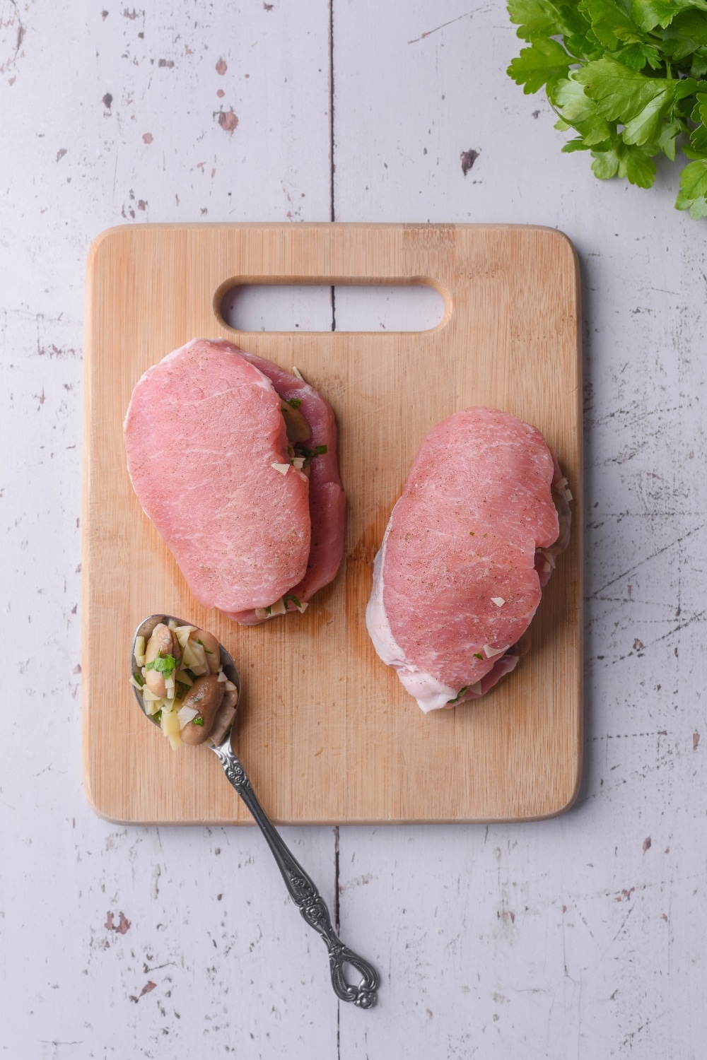 Raw stuffed pork chops on a wooden cutting board. A spoon with mushrooms and swiss cheese stuffing rests on the board.