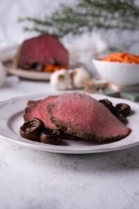 Two slices of medium rare eye of round steak with mushrooms on a white plate. In the back is a bowl of baby carrots and the rest of the steak.