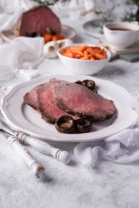 Three slices of medium rare eye of round steak with mushrooms on a white plate. In the back is a small bowl of baby carrots, gravy, and the rest of the steak.