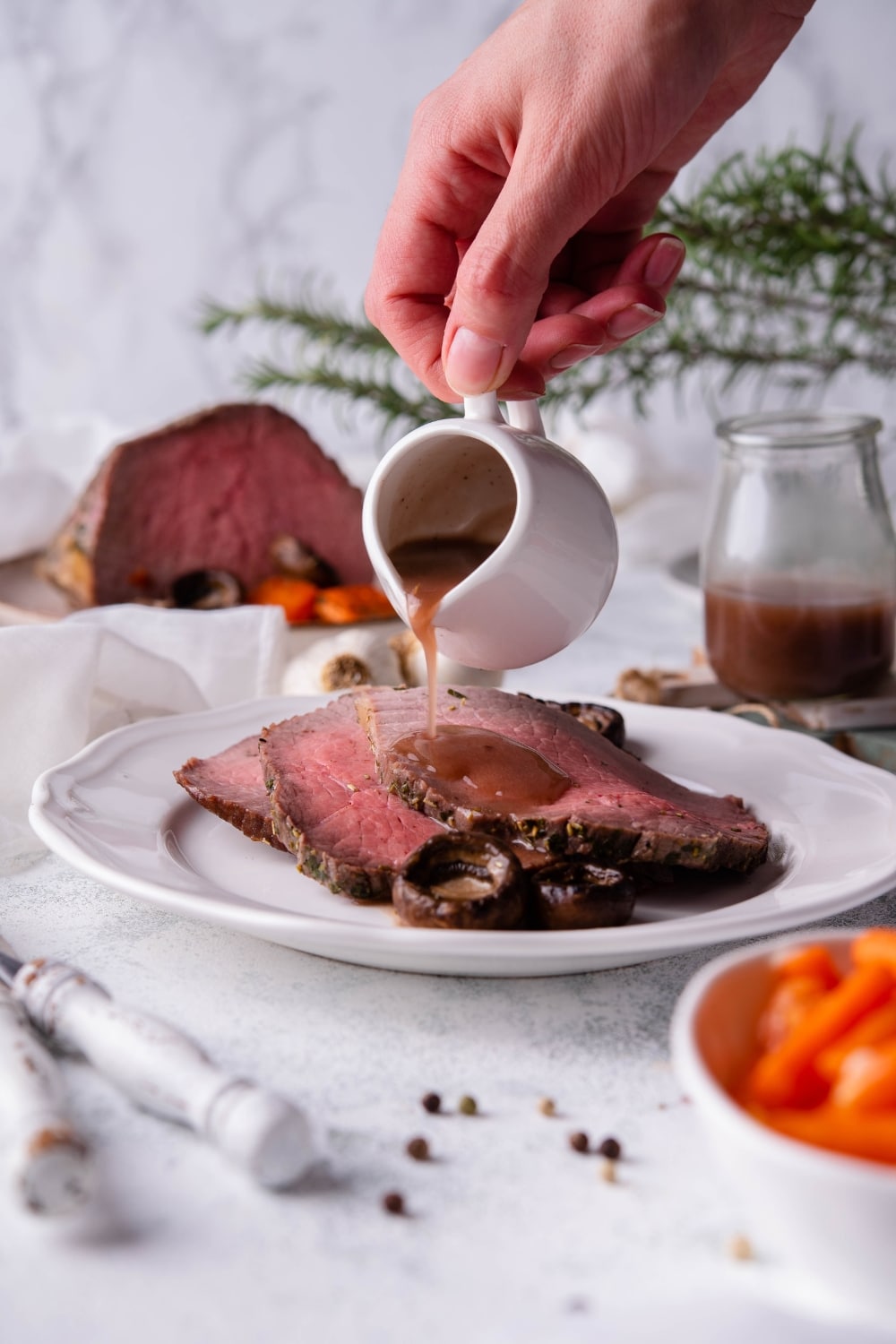 Two slices of medium rare eye of round steak with mushrooms on a white plate. Gravy is being poured onto the slices from a small white pitcher. In the back is a pitcher of gravy and the rest of the steak, and in the foreground is a bowl of baby carrots.