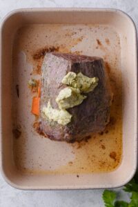 Partially cooked eye of round steak topped with seasoned butter on a ceramic baking tray. Pieces of carrots can be seen under the meat.