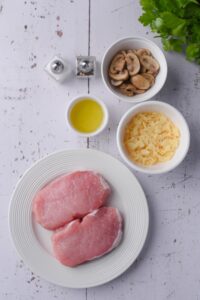 Raw boneless pork chops on a plate next to white bowls of chopped swiss cheese, oil, sliced mushrooms, and salt and pepper.