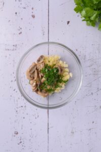 A glass bowl filled with chopped swiss cheese, sliced mushrooms, and chopped parsley.