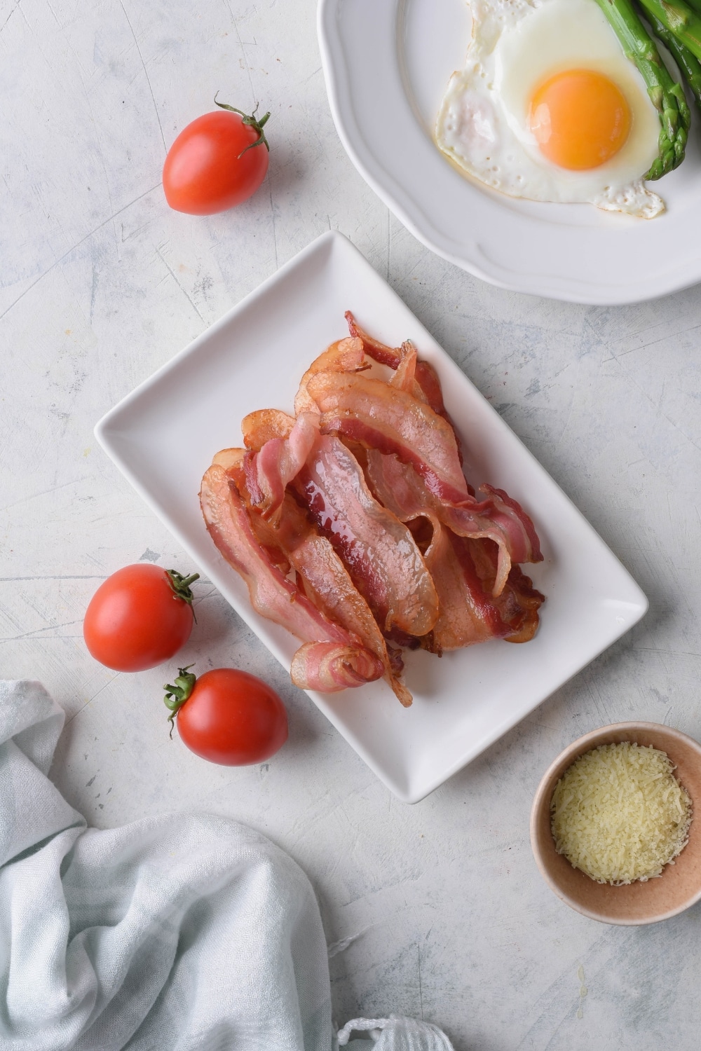 Air fried bacon strips on a rectangle plate. Surrounding it are cherry tomatoes, a small bowl of shredded cheese, and part of a plate of fried eggs and asparagus.