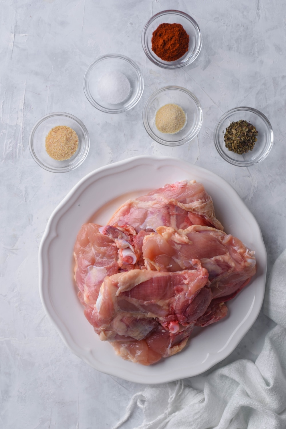 Raw chicken thighs on a plate. Next to it are small glass bowls of smoked paprika, garlic powder, onion powder, oregano, and salt.