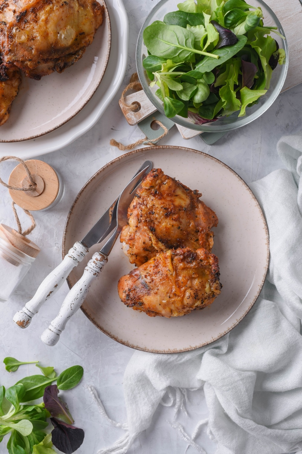 Air fryer chicken thighs on a plate with a knife and fork. Next to it are a bowl of salad and part of a plate of air fried chicken thighs.