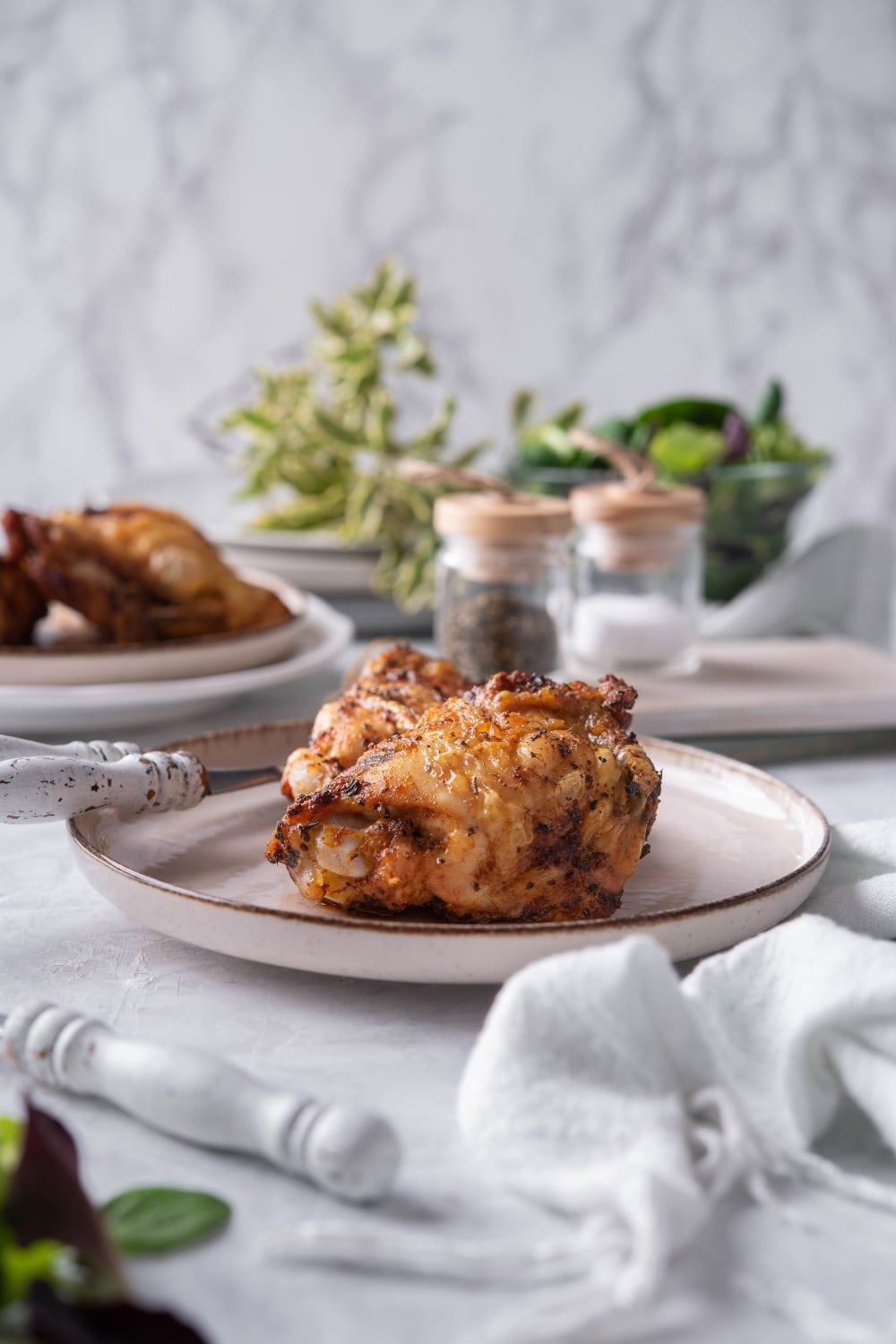 Air fried chicken thighs on a plate with a knife and fork. Behind it is a plate with more air fried chicken thighs and a pair of salt and pepper shakers.