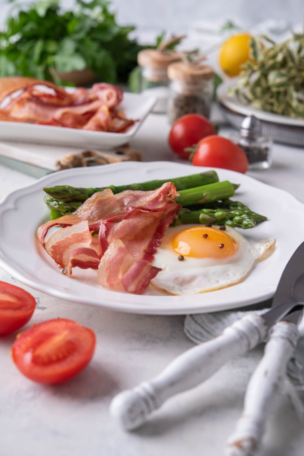 Air fried bacon strips, a sunny side up egg, and cooked asparagus on a white plate. Behind the plate are cherry tomatoes, seasonings, and a plate of more air fried bacon.