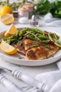 Grilled tilapia filets with sauteed green beans and lemon wedges on a white plate. A fork and knife are next to the plate.