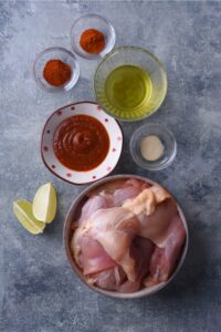 Chicken thighs in a bowl, bbq sauce in a bowl, olive oil in a bowl, cayenne pepper in a bowl, and chili powder in a bowl all on a grey counter.