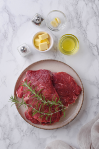 A bowl of butter, a bowl of avocado oil, salt and pepper shakers, and two sirloin steaks on a plate.