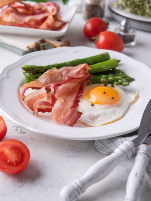 Air fried bacon strips, a sunny side up egg, and cooked asparagus on a white plate.