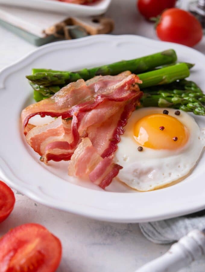 Air fried bacon strips, a sunny side up egg seasoned with pepper, and cooked asparagus on a white plate.