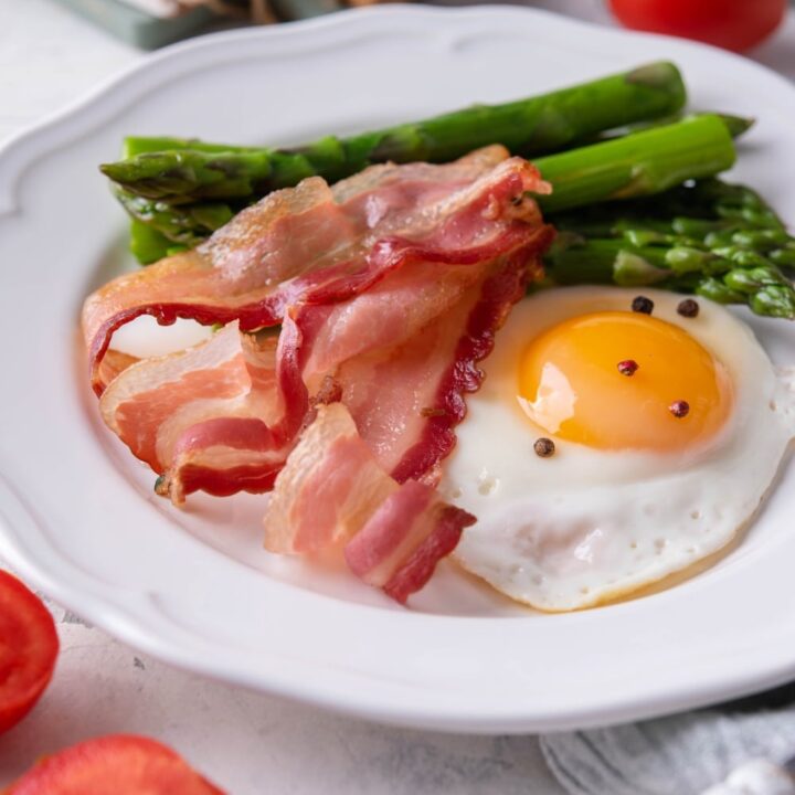 Air fried bacon strips, a sunny side up egg seasoned with pepper, and cooked asparagus on a white plate.