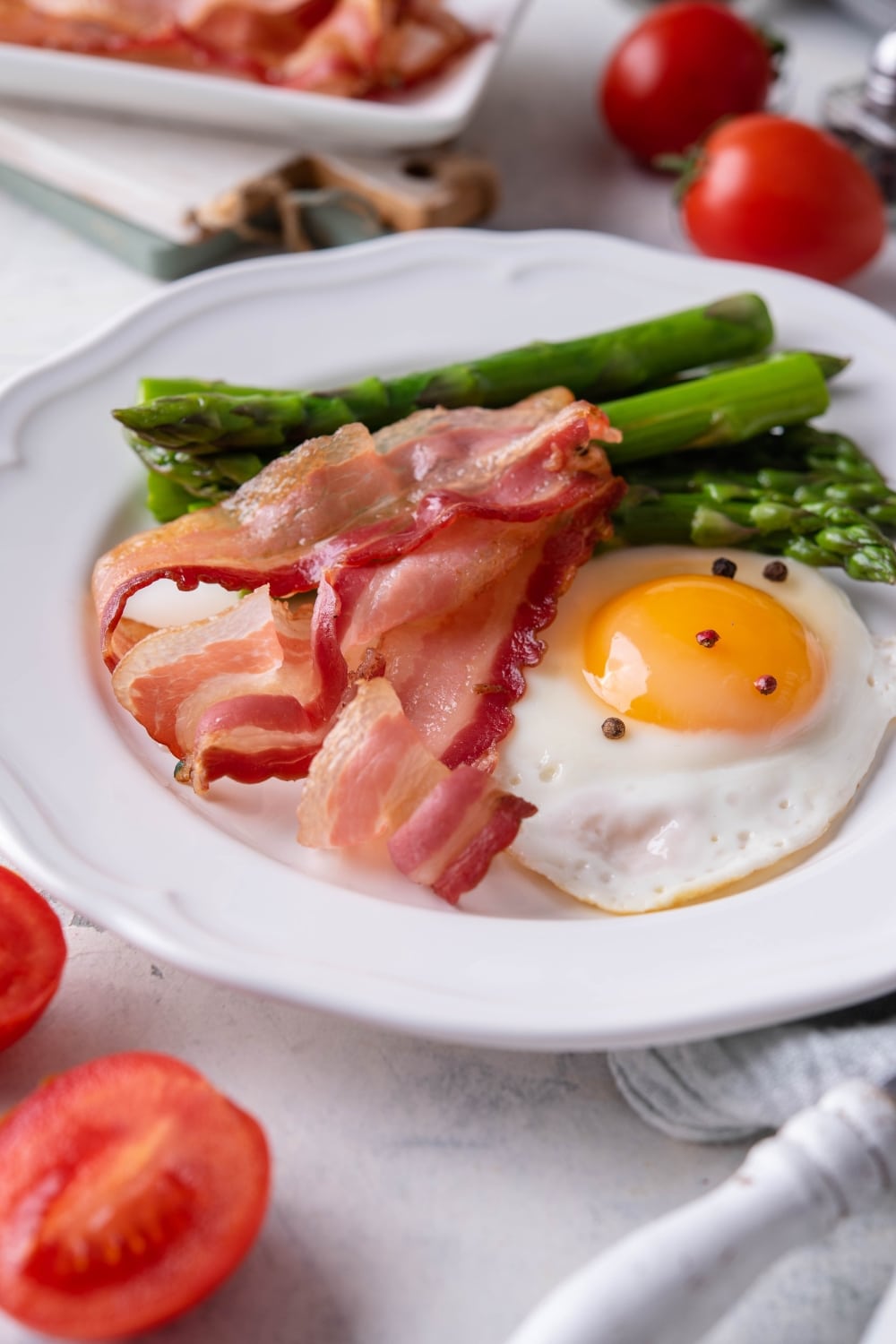 Air fried bacon strips, a sunny side up egg seasoned with pepper, and cooked asparagus on a white plate.