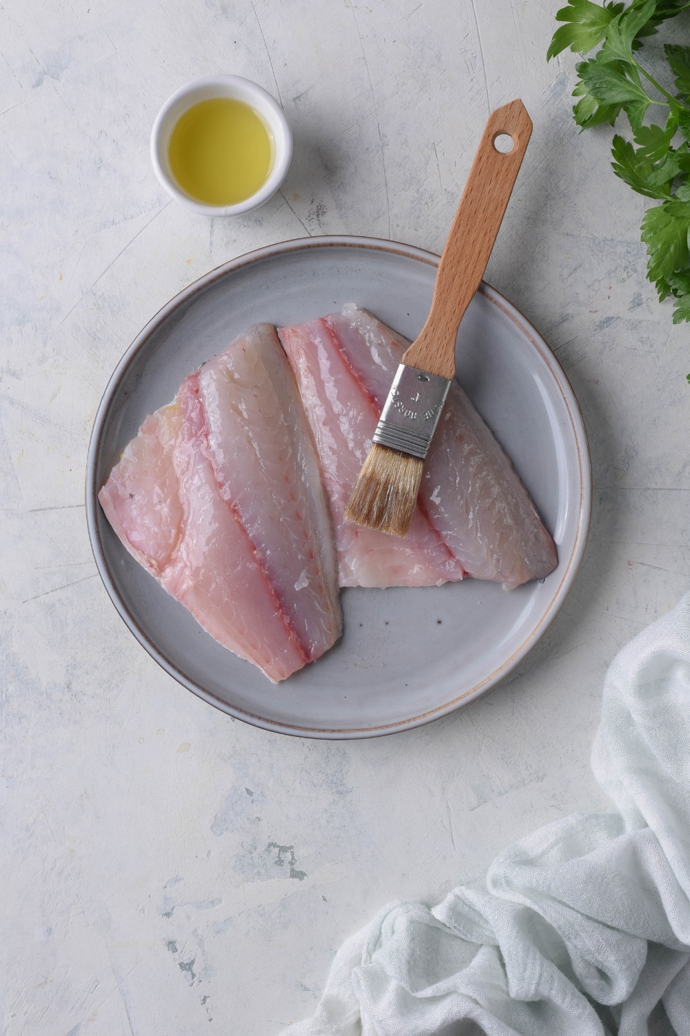 Raw tilapia filets on a plate. A pastry brush dipped in oil sits on top, next to a small saucer of oil.