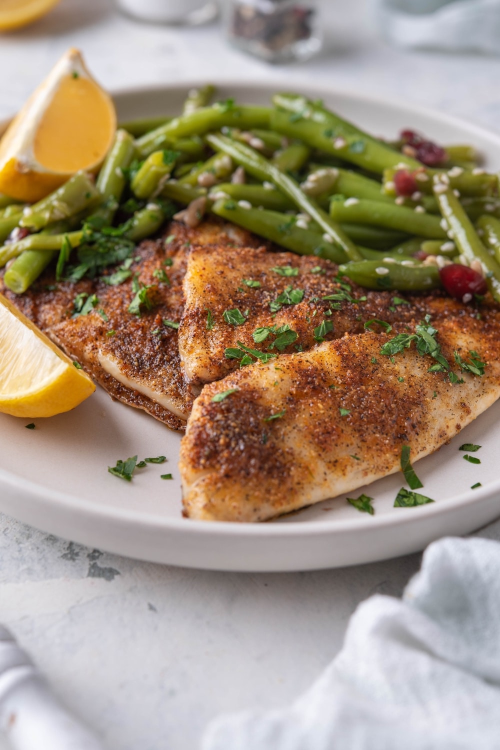 Close up of grilled tilapia filets with sauteed green beans and lemon wedges on a white plate.