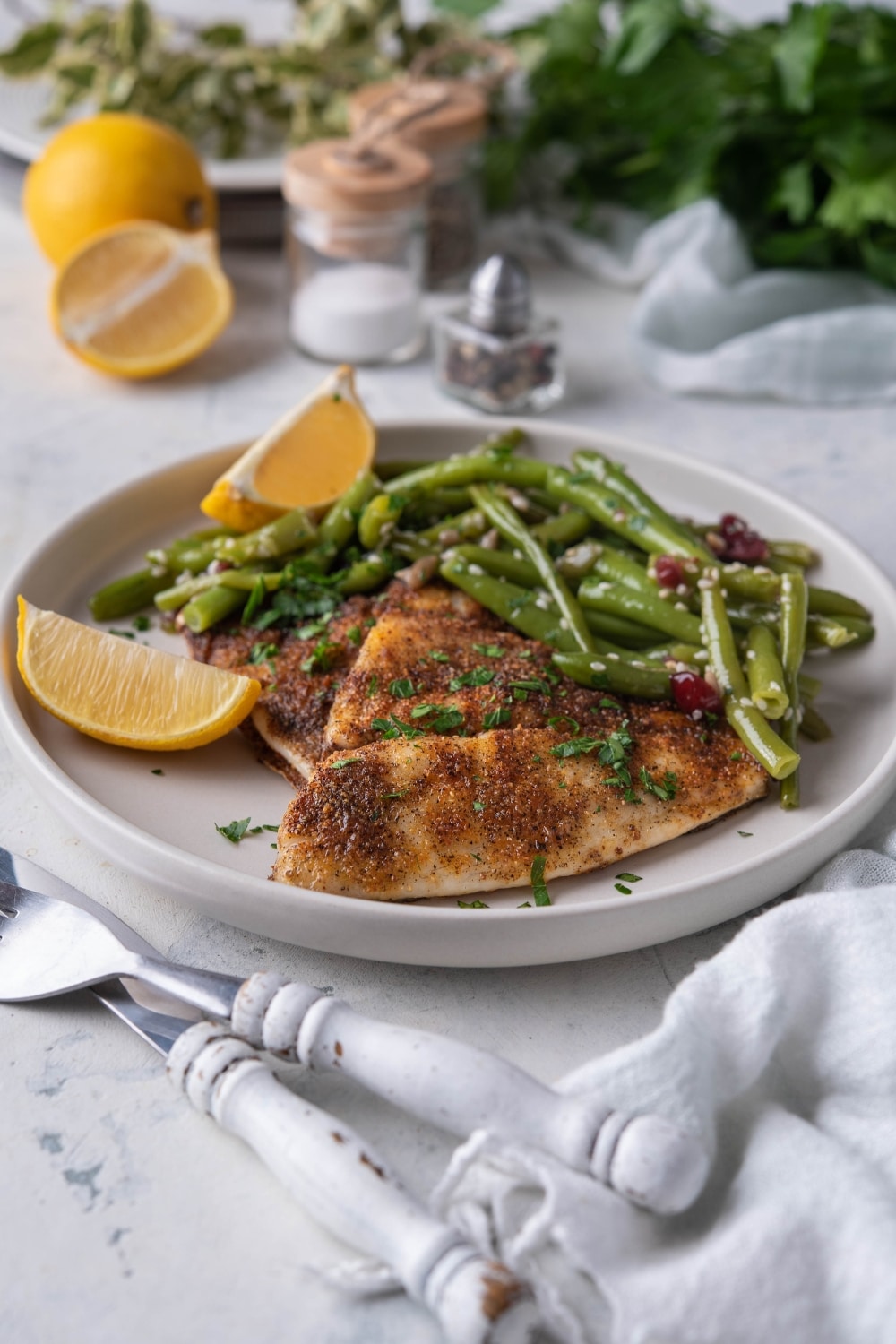 Two grilled tilapia filets with sauteed green beans and lemon wedges on a white plate.