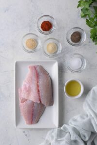 Raw tilapia filets on a rectangle plate. Next to it are small bowls of smoked paprika, onion powder, garlic powder, salt, pepper, and oil.