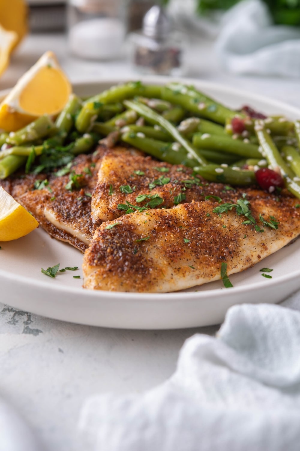 Seasoned grilled tilapia filets with sauteed green beans and lemon wedges on a white plate.