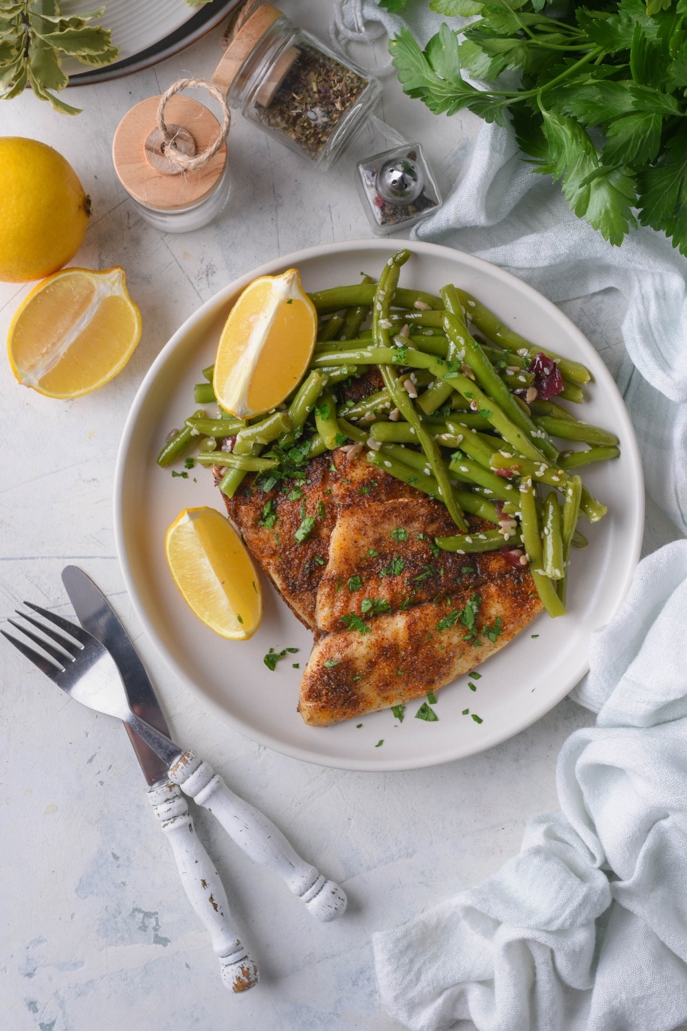 Top view of grilled tilapia filets with sauteed green beans and lemon wedges on a white plate. Surrounding the plate are a fork and knife, tea towel, lemons, and spices.