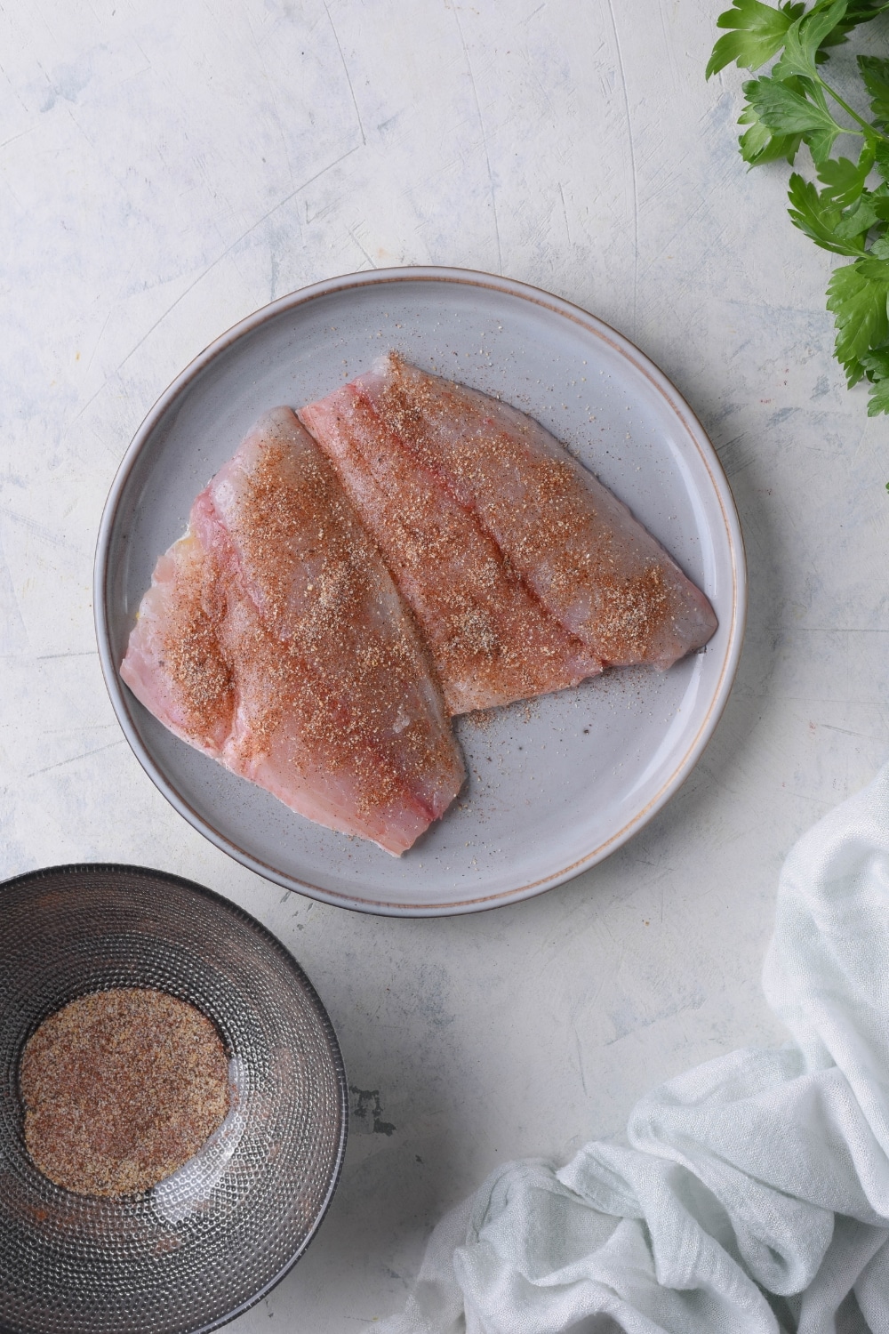 A pair of seasoned raw tilapia filets on a plate. Next to the plate is a bowl of mixed seasonings.