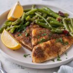 A pair of grilled tilapia filets served with sauteed gren beans and lemon wedges on a white plate.