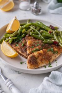 A pair of grilled tilapia filets served with sauteed gren beans and lemon wedges on a white plate.