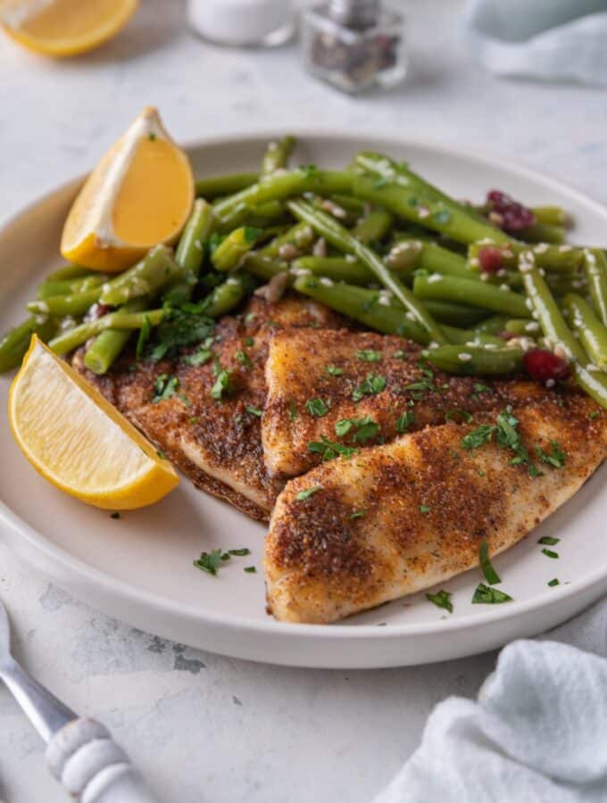 A pair of grilled tilapia filets served with sauteed gren beans and lemon wedges on a white plate.