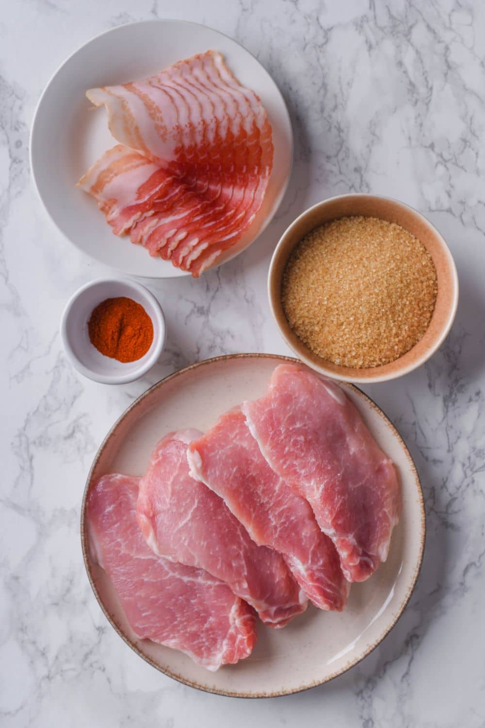 Four raw boneless pork chops on a plate, a plate of bacon, a bowl of brown sugar, and a small bowl of chili powder.