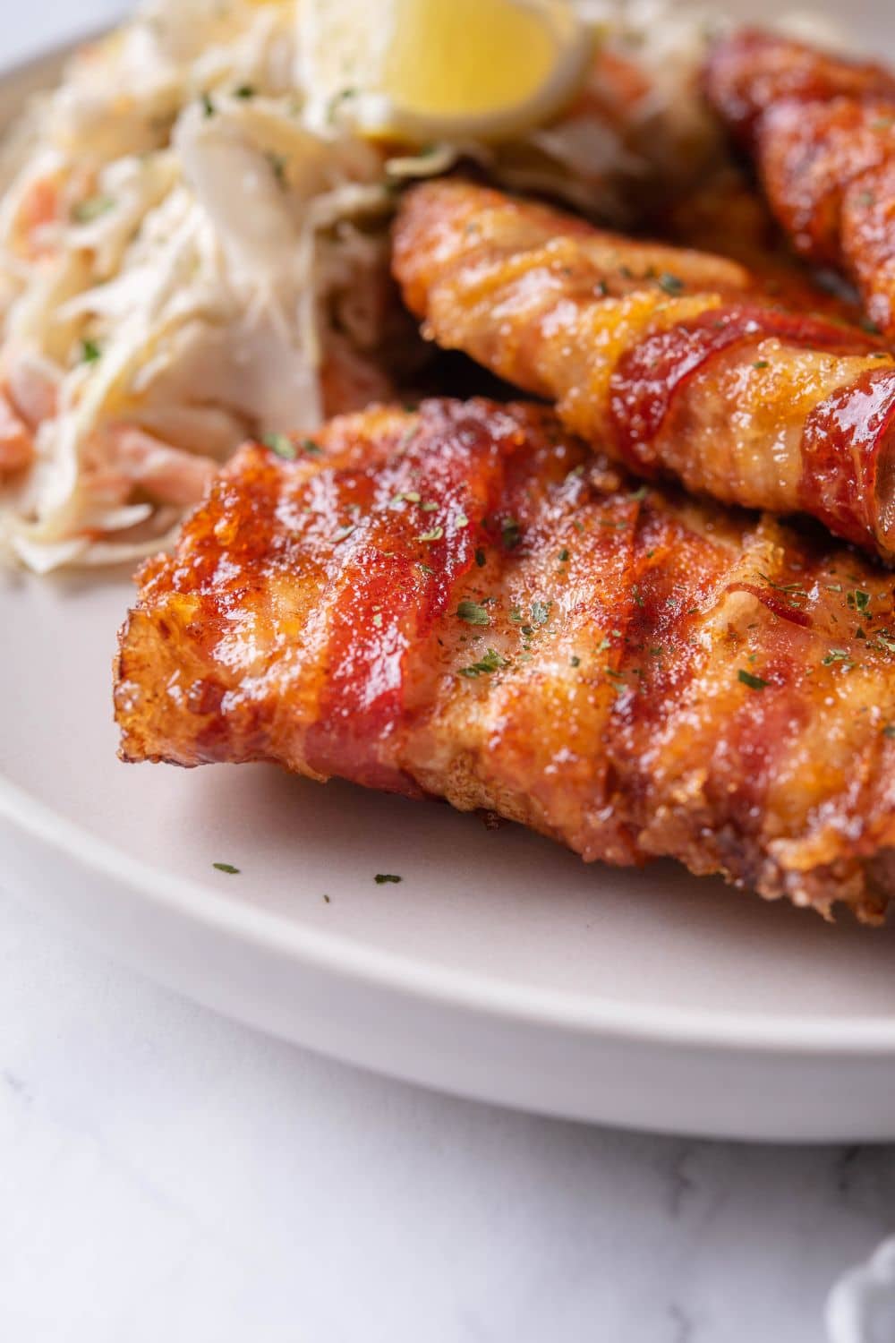 Close up of brown sugar and chili rubbed bacon wrapped pork chops on a plate with slaw and lemon wedges.