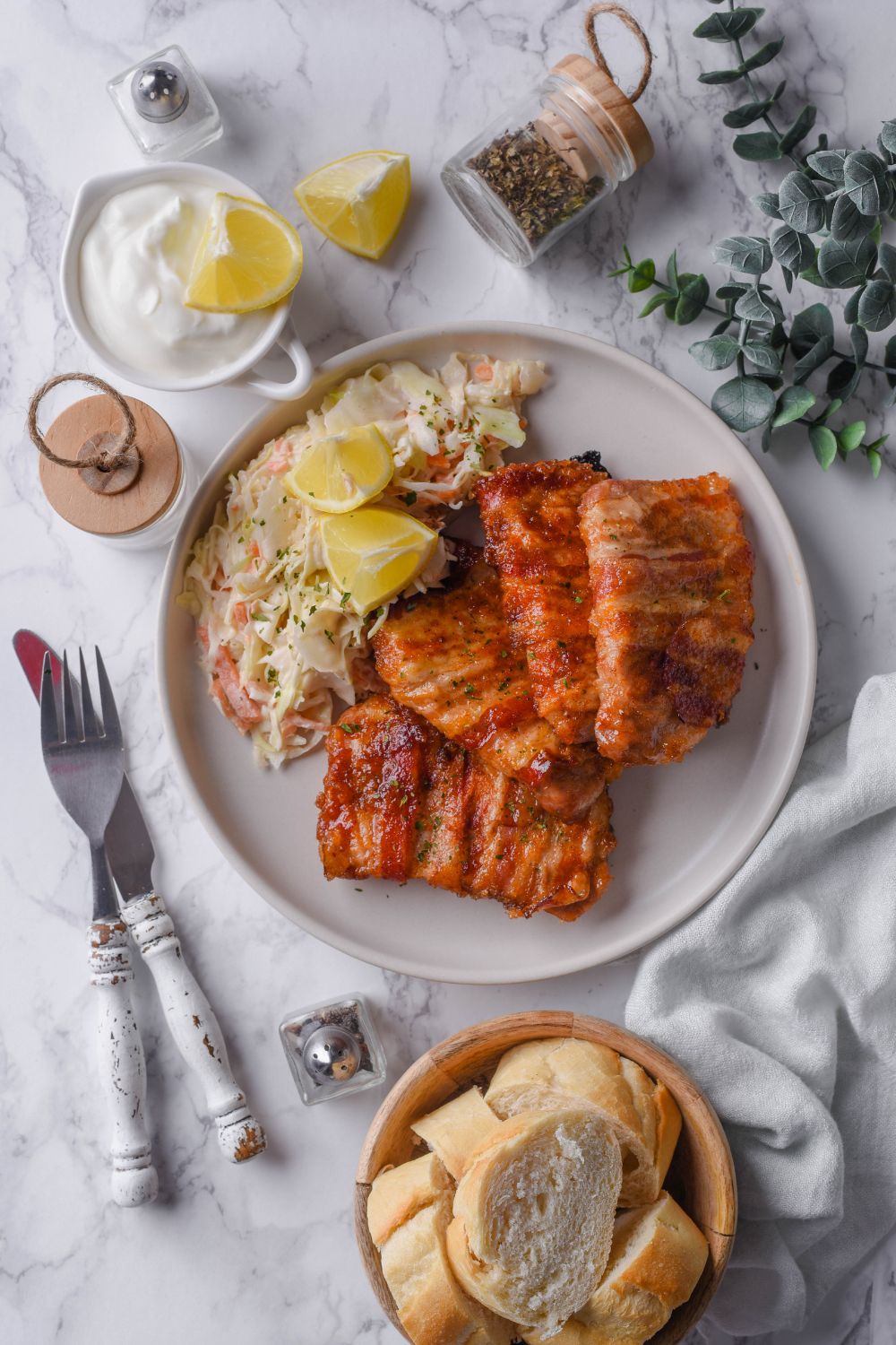 Bacon wrapped pork chops with a side of coleslaw and lemon wedges on a plate, surrounded by a fork and knife, creamy dip, a bowl of bread, and seasonings.