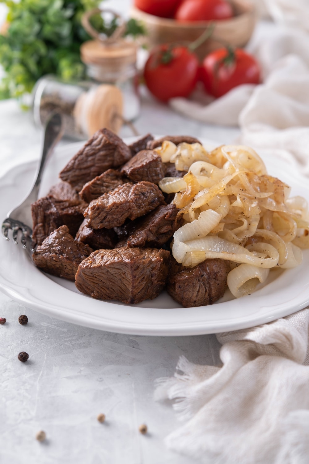 Steak tips and caramelized onions on a white plate with a fork. A bowl of tomatoes and a pair of salt and pepper shakers can be seen in the back.