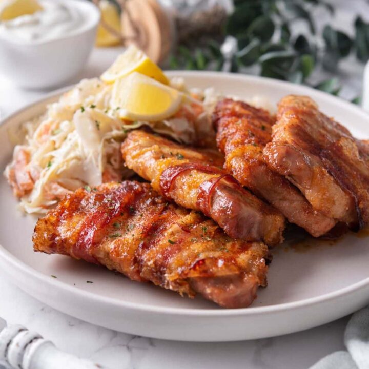 Bacon wrapped pork chops and coleslaw with lemon wedges on a white plate. In the back is a small bowl of creamy dip and a bowl of bread.