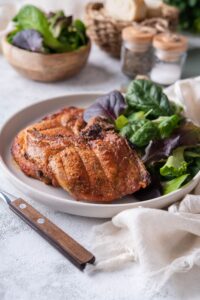 Two broiled bone-in pork chops on a plate with a side of salad greens. In the back are bowls of salad greens and bread slices and salt and pepper shakers.