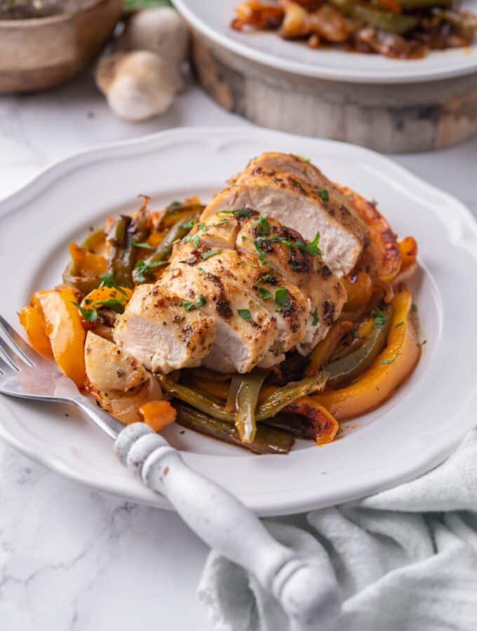 Sliced chicken breast over peppers on a white plate with a fork. Part of a second plate of chicken and peppers is behind.