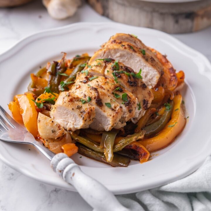 Sliced chicken breast over peppers on a white plate with a fork. Part of a second plate of chicken and peppers is behind.