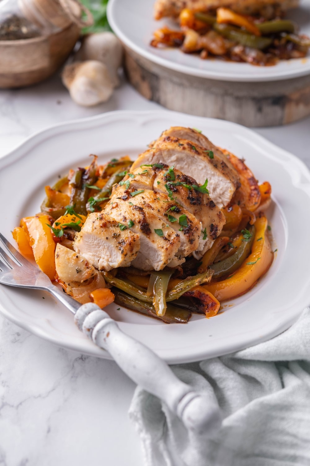 Sliced chicken breast over peppers on a white plate with a fork. Part of a second plate of chicken and peppers is behind.