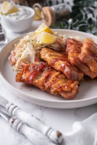 Four bacon wrapped pork chops on a plate with a side of coleslaw and lemon wedges.