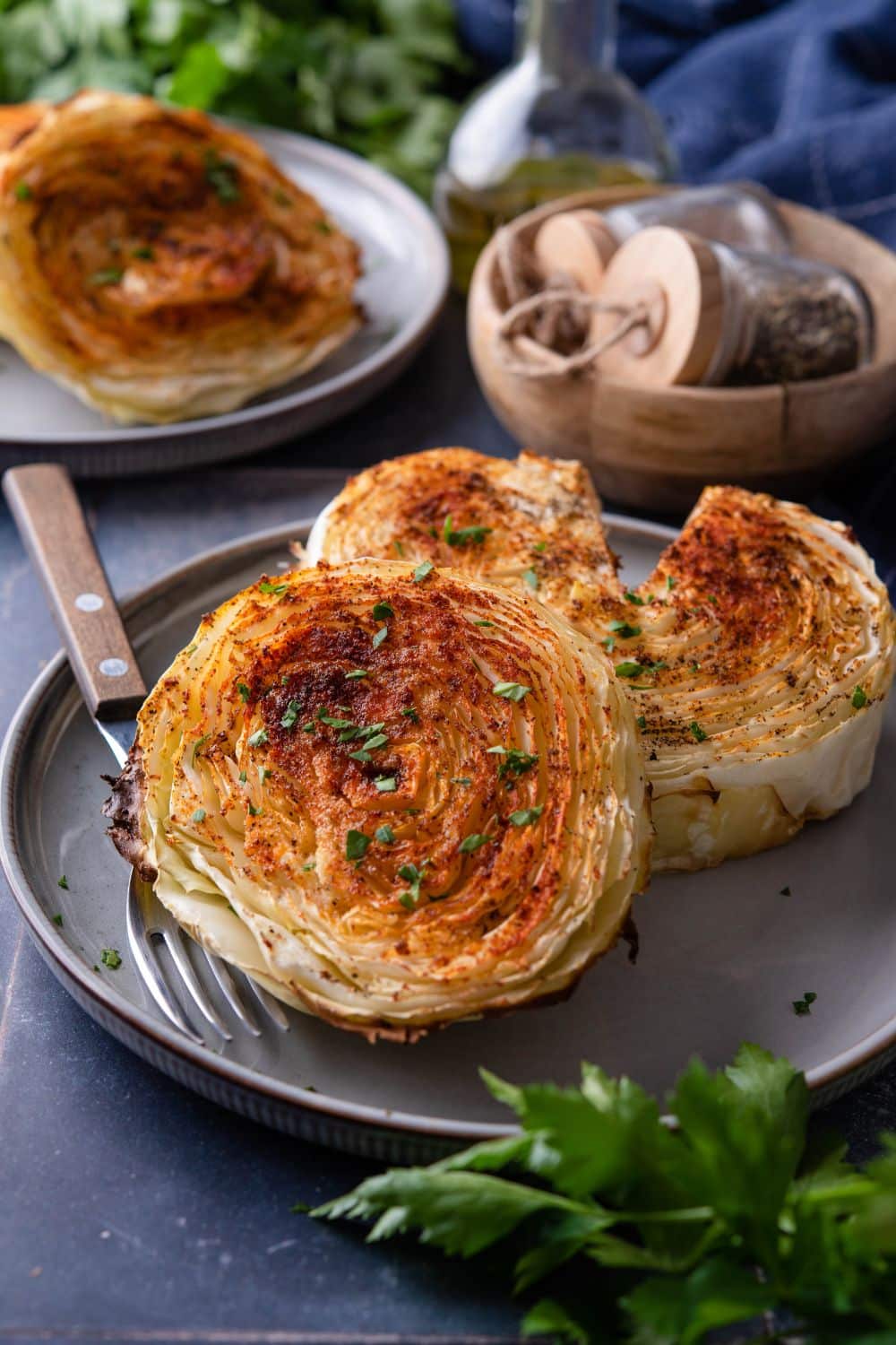 Two roasted cabbage steaks on a plate with a fork. One cabbage steak has a slice taken out. In the back is another plate of cabbage steaks.