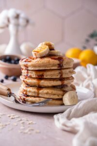 A stack of five oat flour pancakes topped with banana slices and date syrup on a plate with two forks.