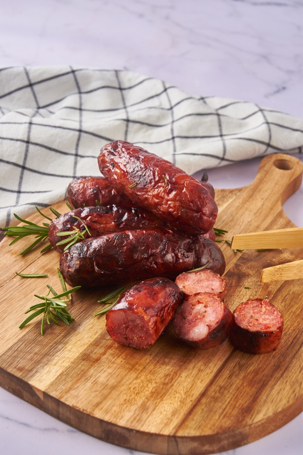 Oven cooked sausages on a wooden serving board. One of the sausages is sliced and a pair of wooden tongs is resting on the board.