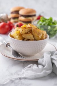 Sauteed chopped potatoes in a white bowl, set on a plate with a fork. Behind it is a bowl of greens and some hamburgers.